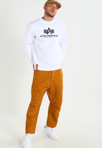 Witte sweatshirt met zwart "ALPHA INDUSTRIES" logo, gecombineerd met oranje utilitybroek en witte sneakers. Casual, sportieve outfit.