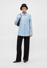 Ανοιχτό μπλε πουκάμισο από τζιν με κουμπιά, με μια μόνο τσέπη στο στήθος, σε συνδυασμό με μαύρο παντελόνι σε φάρδος και μαύρες στυλιζαρισμένες μούλες, ολοκληρωμένο με ένα μαύρο καπέλο τύπου bucket.