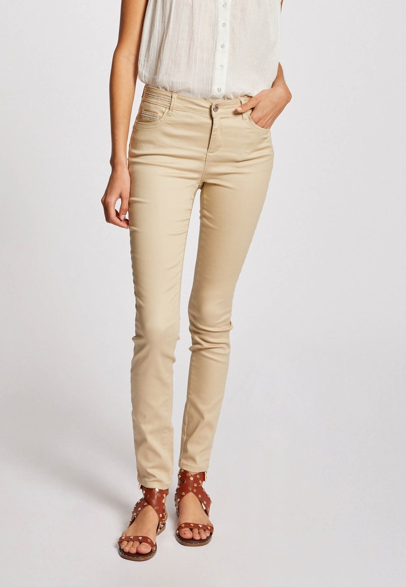 Beige slim-fit broek met een gladde textuur en een klassiek vijf-pocket ontwerp, gecombineerd met bruine sandalen met bandjes en decoratieve studs.