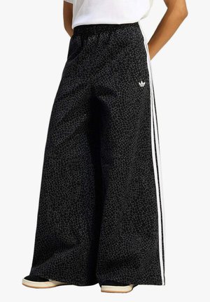 Persona che indossa pantaloni neri a gamba larga Adidas con un motivo crepato sottile, strisce laterali bianche e scarpe da ginnastica nere con suole bianche.