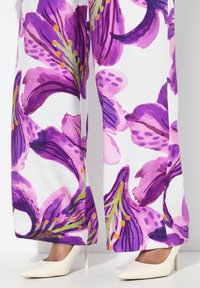 Los pantalones de pierna ancha con estampado de flores presentan vibrantes flores moradas sobre un fondo blanco, combinados con unos tacones blancos de punta.