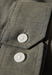 Textil gris con una textura fina, que presenta dos botones redondos blancos y un cuello puntiagudo. Las costuras visibles añaden detalle a la tela.
