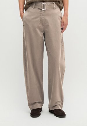 Personne portant un pantalon beige à jambes larges avec ceinture et poches latérales, associée à des chaussures marron foncé, debout devant un fond uni.