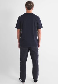 Homme de dos portant un t-shirt à manches courtes bleu marine, un pantalon cargo sombre et des baskets noires avec semelles blanches sur un fond uni.