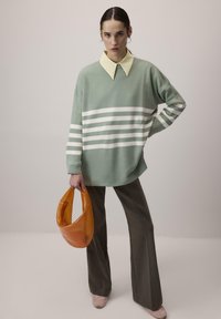 Mintgroene trui met witte strepen, gele kraag, gecombineerd met donkerbruin flared broek. Houdt een oranje glanzende handtas vast.