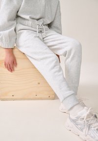 Hellgrauer Hoodie und passendes Sweatpants mit elastischem Bund und Bündchen, getragen mit weißen und silbernen Sneakers, auf einer Holzoberfläche.