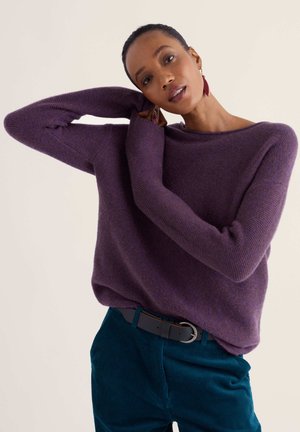 Femme portant un pull violet et un pantalon en velours côtelé bleu-vert avec une ceinture noire, ajustant sa boucle d'oreille avec un bras levé derrière la tête.