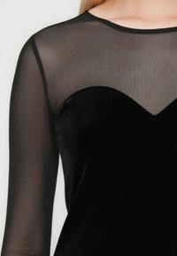 Zwart fluwelen jurk met sheer mesh lange mouwen en bovenstuk, voorzien van een sweetheart halslijn detail.