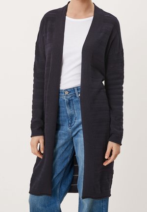 Femme portant un long cardigan noir à ouverture avant avec des rayures texturées sur un t-shirt blanc et un jean bleu, debout devant un fond uni.