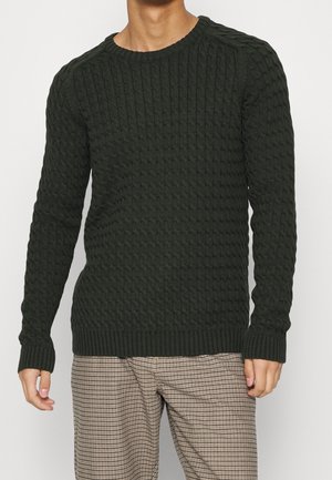 Man die een donkergroen kabelgebreide trui en beige geruite broek draagt, staand met ontspannen armen langs zijn zij.