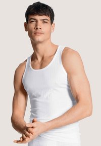 Witte tanktop met een geribbelde textuur en brede schouderbanden, gedragen door een model die met gevouwen handen staat.
