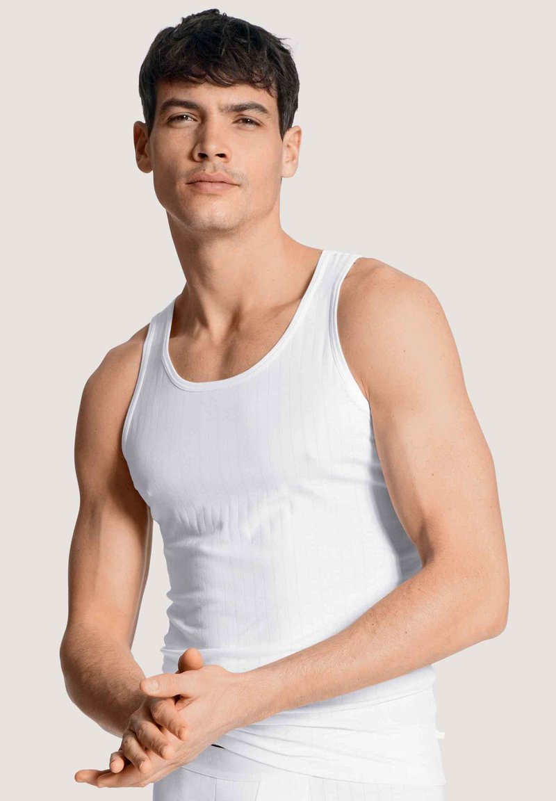Witte tanktop met een geribbelde textuur en brede schouderbanden, gedragen door een model die met gevouwen handen staat.