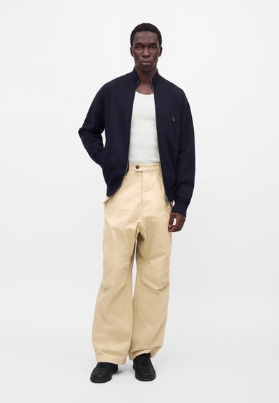 Homme portant un pantalon beige à jambes larges, un débardeur blanc et une veste zippée bleu marine, debout avec une main dans la poche sur fond blanc.