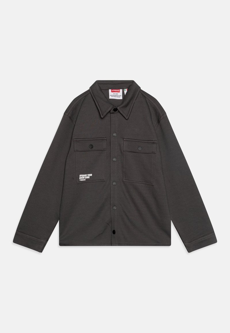 Veste-chemise gris foncé à boutons, manches longues, deux poches poitrine avec boutons, et un petit texte blanc imprimé sur la poche gauche.