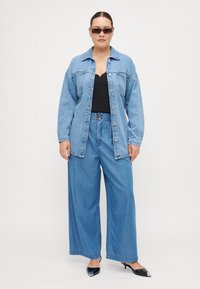 Light blue denim jacket with button-up front, paired with wide-leg light blue denim pants. Black top underneath and pointed black shoes.