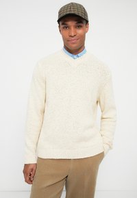 Homem a usar um suéter de malha bege sobre uma camisa de colarinho azul, calças de veludo cotelê castanhas e uma boina xadrez, de pé com uma mão no bolso.