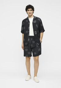 Jeune homme portant une chemise noire à manches courtes assortie et un short avec des motifs abstraits gris clair, un t-shirt blanc et des chaussures blanches sans lacets.