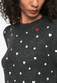Ciemnoszara sweter z dzianiny, z rozrzuconymi białymi sercami i jednym czerwonym sercem. Okrągły dekolt z miękką teksturą. Zbliżenie.