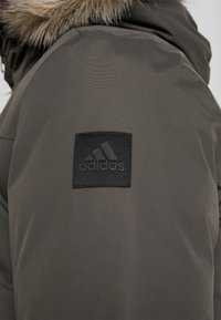 Veste d'hiver vert foncé avec une capuche bordée de fourrure et un patch logo Adidas carré noir sur la manche supérieure.