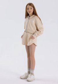 Sudadera con capucha beige con cremallera, pantalones cortos a juego y zapatillas blancas. Incluye logo bordado y puños acanalados. Textura de tela suave.