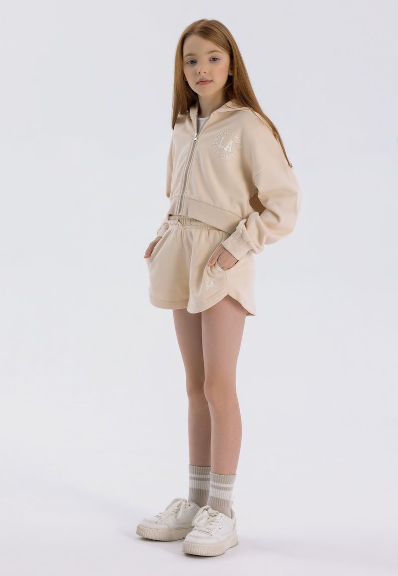 Sudadera con capucha beige con cremallera, pantalones cortos a juego y zapatillas blancas. Incluye logo bordado y puños acanalados. Textura de tela suave.