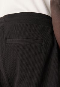 Pantaloni neri in tessuto testurizzato con vita a coste e tasca posteriore. Il tessuto presenta un motivo a griglia, offrendo una texture visiva distintiva.