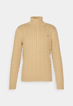 Suéter de cuello alto color beige claro con diseño de trenzas, puños y dobladillo acanalados. Presenta un pequeño logo azul en el pecho.