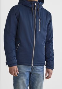 Giubbotto puffo blu navy con cappuccio, chiusura con zip e tasche laterali. Presenta coulisse regolabili e una superficie liscia, resistente all'acqua.
