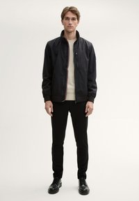 Zwarte zip-up jack met een hoge kraag, witte accenten, gedragen over een licht crèmekleurige trui, gecombineerd met zwarte broek en zwarte schoenen.