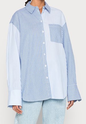Person wearing oversized, long-sleeve button-up shirt with mixed blue and white vertical stripes and a front pocket, paired with light blue jeans.