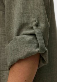 Chemise en tissu texturé vert avec des manches retroussées et un détail de bouton. Tissage de tissu visible et variation tonale subtile.