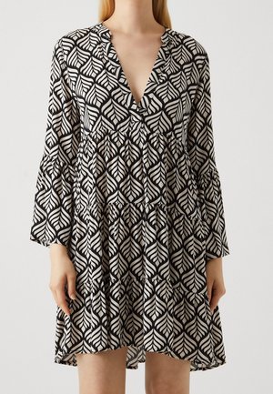 Woman wearing a black and white leaf-patterned dress with long bell sleeves and a deep V-neckline, standing against a plain background.