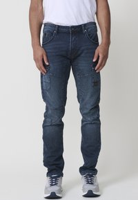 Pantalones cargo de mezclilla en azul oscuro con múltiples bolsillos, un ajuste slim, áreas de rodilla reforzadas y una textura suave. Camiseta blanca y zapatillas grises.