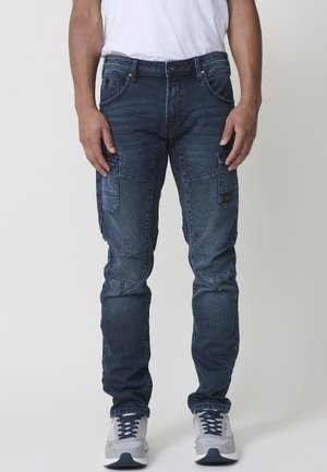 Pantalones cargo de mezclilla en azul oscuro con múltiples bolsillos, un ajuste slim, áreas de rodilla reforzadas y una textura suave. Camiseta blanca y zapatillas grises.