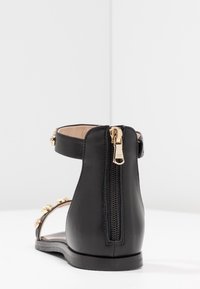 Sandalo in pelle nera con cinturino alla caviglia, hardware dorato, zip sul retro e borchie decorative sui lati, sole piatto.