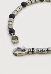 Ασημένιο κολιέ από μέταλλο με μαύρες χάντρες, υφασμένα ασημένια στοιχεία και κούμπωμα με ανάγλυφη τη λέξη «GUESS», παρουσιάζοντας ποικιλία σχημάτων και φινιρισμάτων.