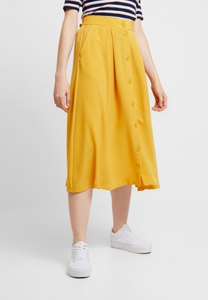 Kvinde iført en midi-nederdel i sennepsgul med knapper foran og lommer, kombineret med en stribet top og hvide sneakers.