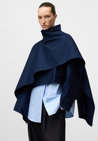 Cappa blu navale con colletto alto, design a strati e dettaglio con bottoni. Sotto, una camicia azzurro chiaro con colletto classico e chiusura a bottoni.