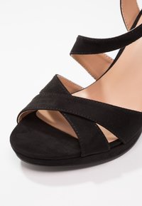 Sandale compensée en suede noir avec des sangles croisées et une semelle intérieure nude lisse. La semelle est arrondie, offrant une texture douce.