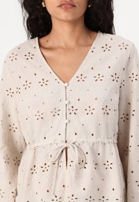 Light beige top with floral eyelet pattern, V-neckline, button details, and adjustable waist tie. Long sleeves with gathered cuffs.