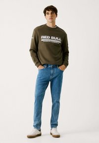 Olivgrön sweatshirt med "RED BULL"-logotyp; kombinerad med ljusblå jeans. Modellen bär vita sneakers med brun sula.