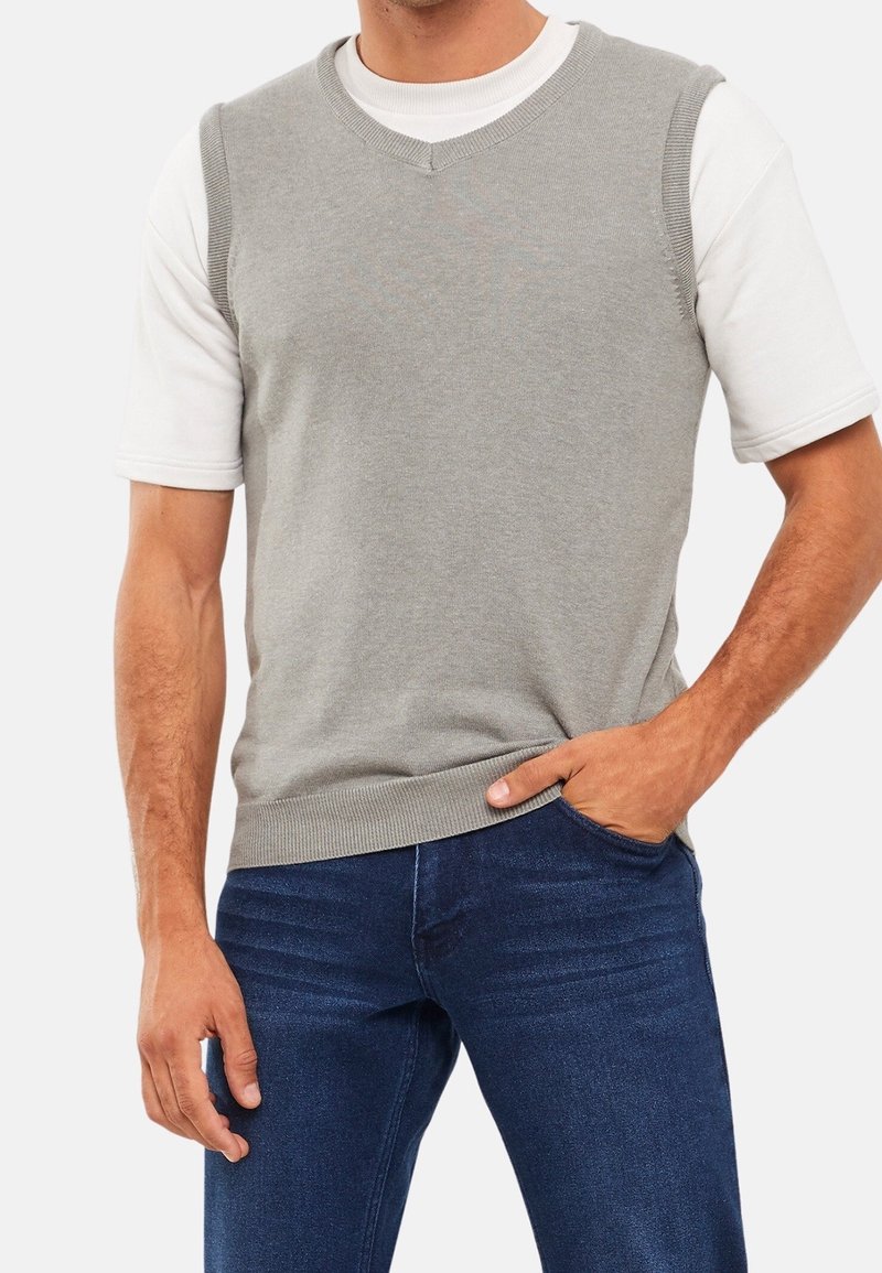 Homme portant un gilet sans manches gris clair sur une chemise blanche à manches courtes, jean bleu foncé, une main dans la poche.