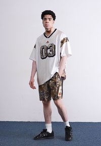 Camisa de jersey branca de mangas curtas com detalhes em preto e camuflagem, combinada com calções de camuflagem correspondentes. Ténis pretos e meias brancas.