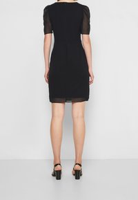 Černé šaty s krátkými průsvitnými nabíranými rukávy, přiléhavým střihem a zapínáním na zip na zádech. Nositelná s černými sandály na vysokém podpatku.