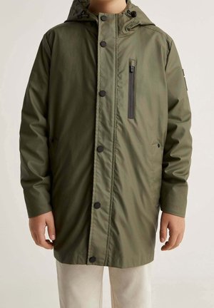 Personne portant un imperméable à capuche vert olive avec des boutons-pression noirs et une poche poitrine verticale zippée, pantalon beige visible sous l'ourlet du manteau.