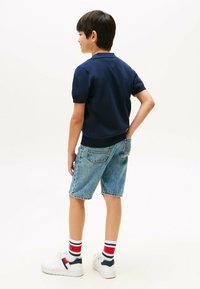 Granatowa koszulka polo z krótkim rękawem, niebieskie szorty dżinsowe, białe sneakersy z granatowymi i czerwonymi akcentami, skarpetki w czerwono-białe paski; postawa stojąca.