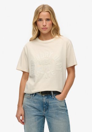 T-shirt à manches courtes de couleur crème fabriqué en coton doux, avec un design de texte en relief et une coupe décontractée. Associé à un jean bleu clair.