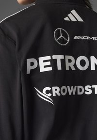 Schwarze Sportjacke mit großen weißen Logos "PETRONAS" und "CROWDSTRIKE", sowie Mercedes- und Adidas-Symbolen in kontrastierenden Farben.