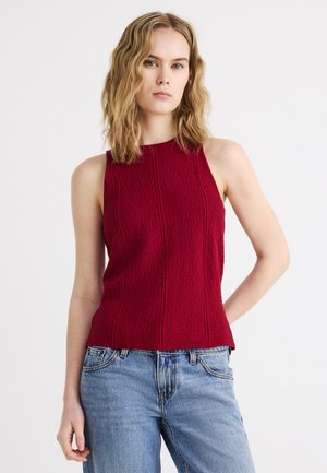Vrouw met schouderlang golvend blond haar, gekleed in een mouwloze, gestructureerde rode top en lichtblauwe spijkerbroek, staand tegen een effen witte achtergrond.