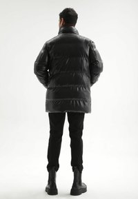 Zwarte gewatteerde jas met hoge kraag, glanzende textuur, horizontaal doorgestikt patroon, gedragen over zwarte broek en stevige zwarte laarzen.